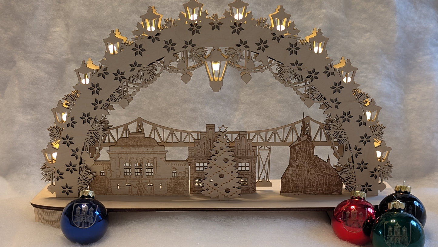 „Holz-Schwibbogen mit filigranen Motiven aus Rendsburg, darunter die Eisenbahn-Hochbrücke, das Landestheater, das alte Rathaus und die St. Marienkirche. Gefertigt aus hellem Birkensperrholz und von innen beleuchtet.“