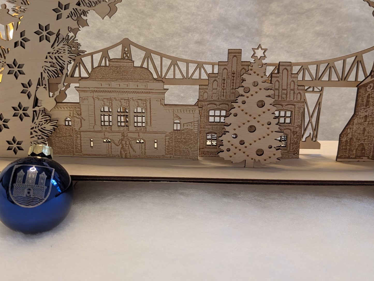 „Holz-Schwibbogen mit filigranen Motiven aus Rendsburg, darunter die Eisenbahn-Hochbrücke, das Landestheater, das alte Rathaus und die St. Marienkirche. Gefertigt aus hellem Birkensperrholz und von innen beleuchtet.“