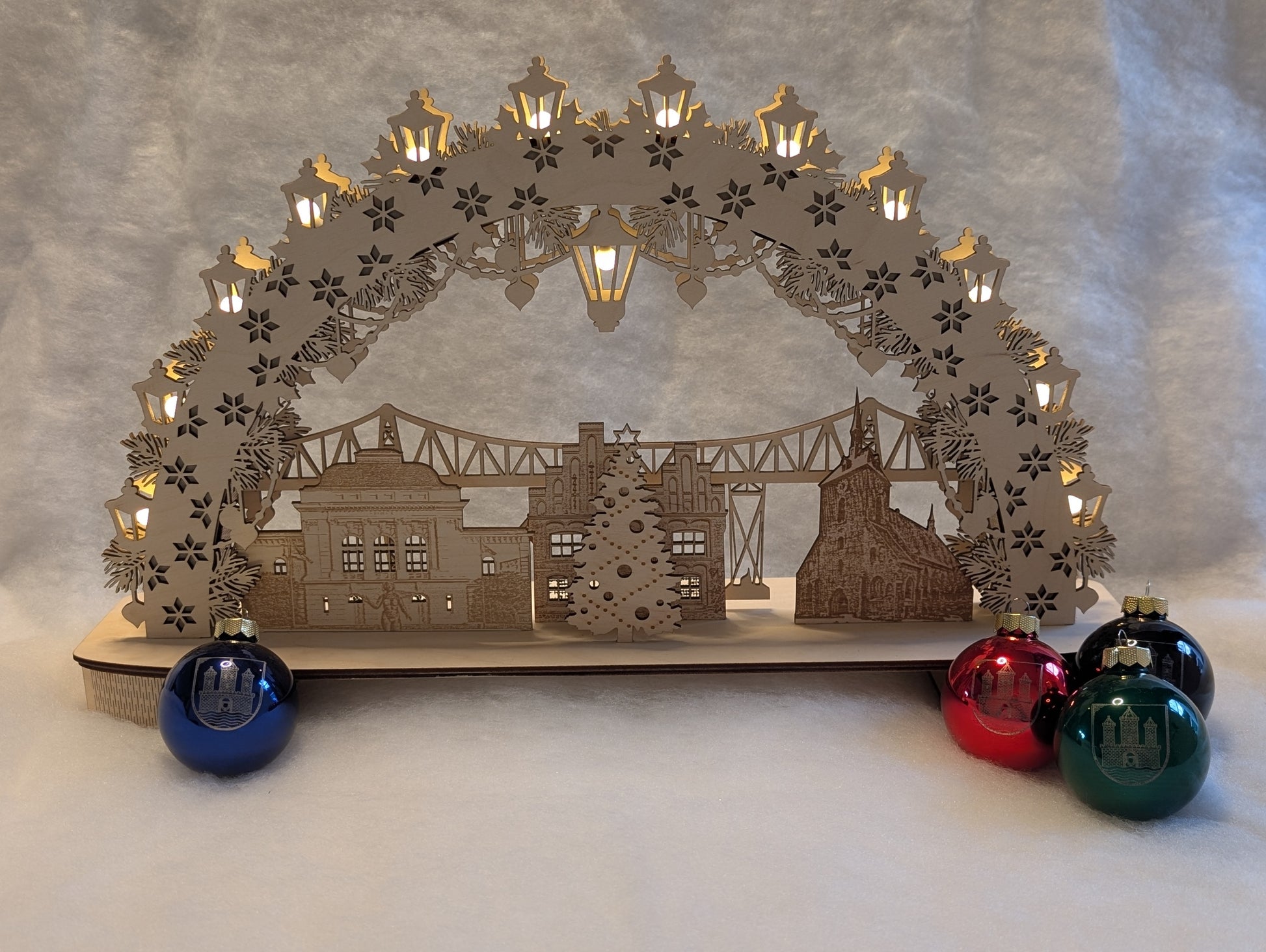 „Holz-Schwibbogen mit filigranen Motiven aus Rendsburg, darunter die Eisenbahn-Hochbrücke, das Landestheater, das alte Rathaus und die St. Marienkirche. Gefertigt aus hellem Birkensperrholz und von innen beleuchtet.“