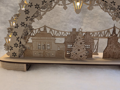 „Holz-Schwibbogen mit filigranen Motiven aus Rendsburg, darunter die Eisenbahn-Hochbrücke, das Landestheater, das alte Rathaus und die St. Marienkirche. Gefertigt aus hellem Birkensperrholz und von innen beleuchtet.“