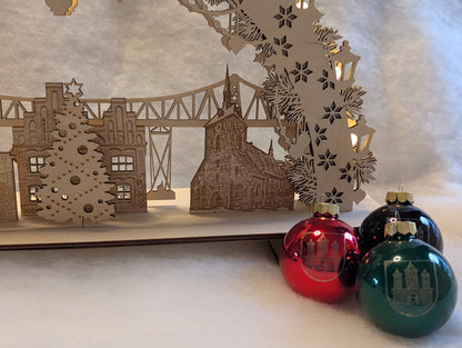 „Holz-Schwibbogen mit filigranen Motiven aus Rendsburg, darunter die Eisenbahn-Hochbrücke, das Landestheater, das alte Rathaus und die St. Marienkirche. Gefertigt aus hellem Birkensperrholz und von innen beleuchtet.“