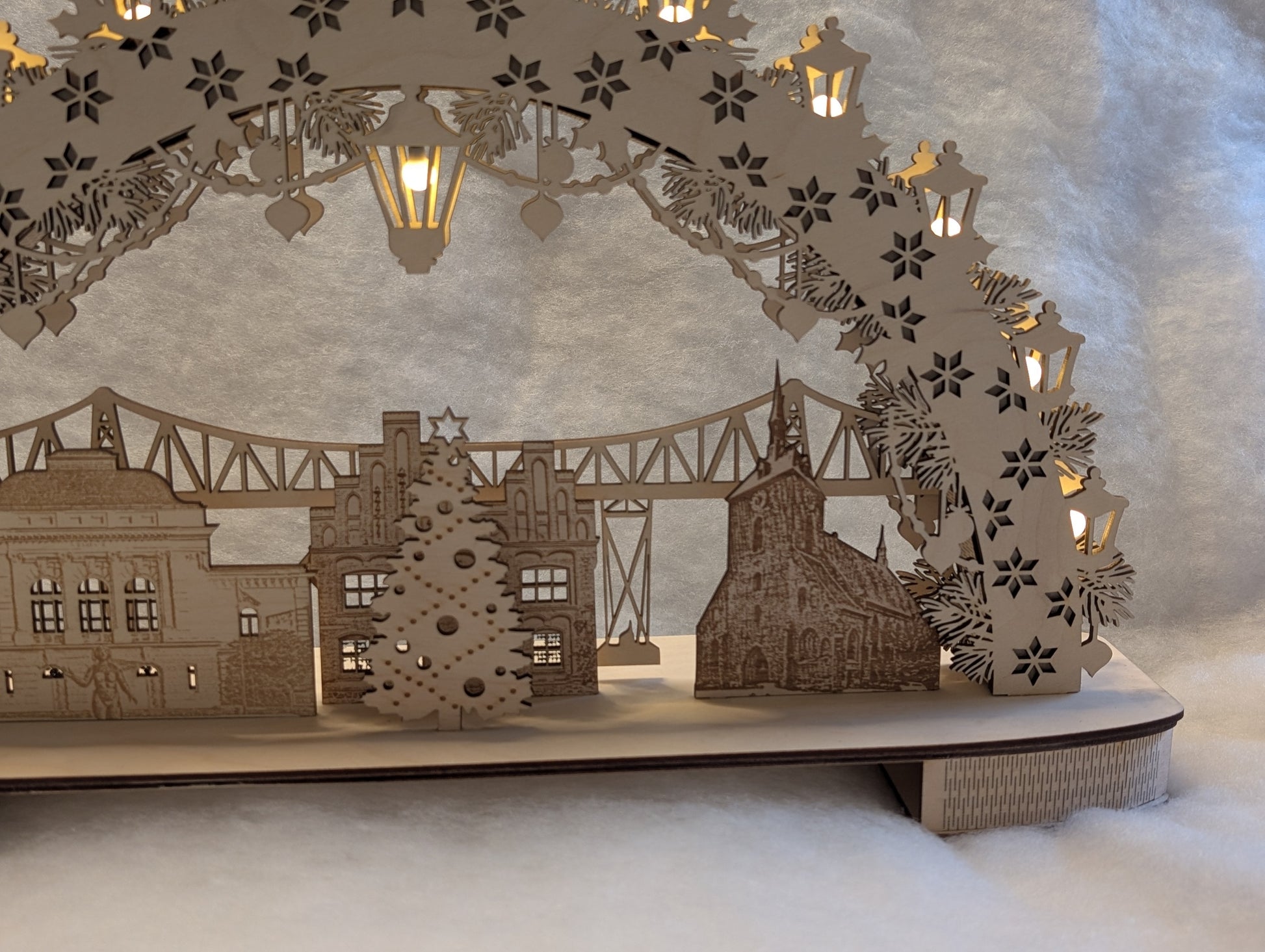 „Holz-Schwibbogen mit filigranen Motiven aus Rendsburg, darunter die Eisenbahn-Hochbrücke, das Landestheater, das alte Rathaus und die St. Marienkirche. Gefertigt aus hellem Birkensperrholz und von innen beleuchtet.“