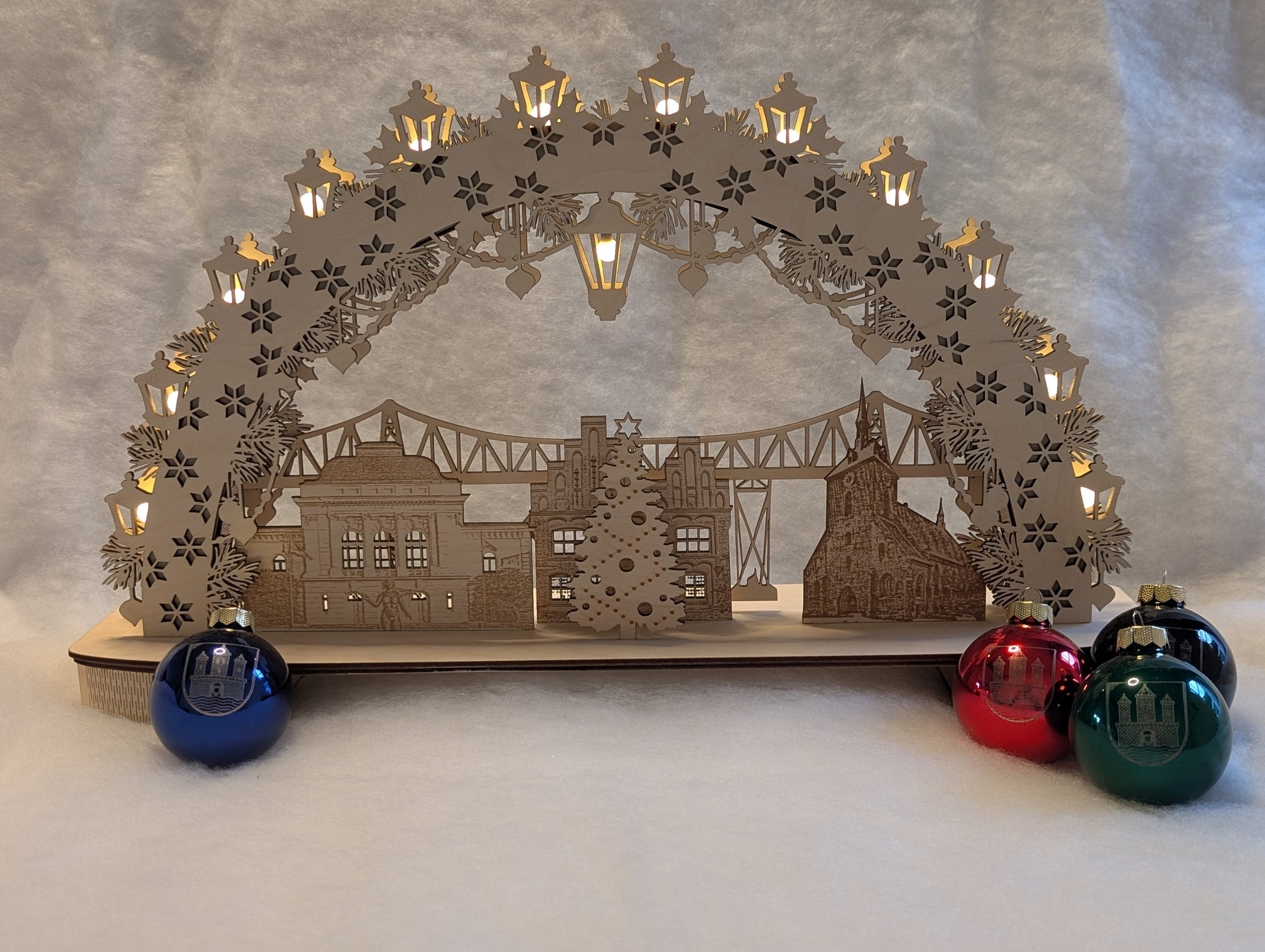 „Holz-Schwibbogen mit filigranen Motiven aus Rendsburg, darunter die Eisenbahn-Hochbrücke, das Landestheater, das alte Rathaus und die St. Marienkirche. Gefertigt aus hellem Birkensperrholz und von innen beleuchtet.“