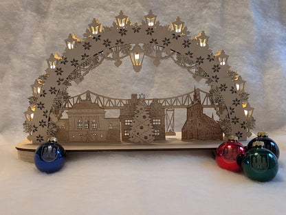 „Holz-Schwibbogen mit filigranen Motiven aus Rendsburg, darunter die Eisenbahn-Hochbrücke, das Landestheater, das alte Rathaus und die St. Marienkirche. Gefertigt aus hellem Birkensperrholz und von innen beleuchtet.“