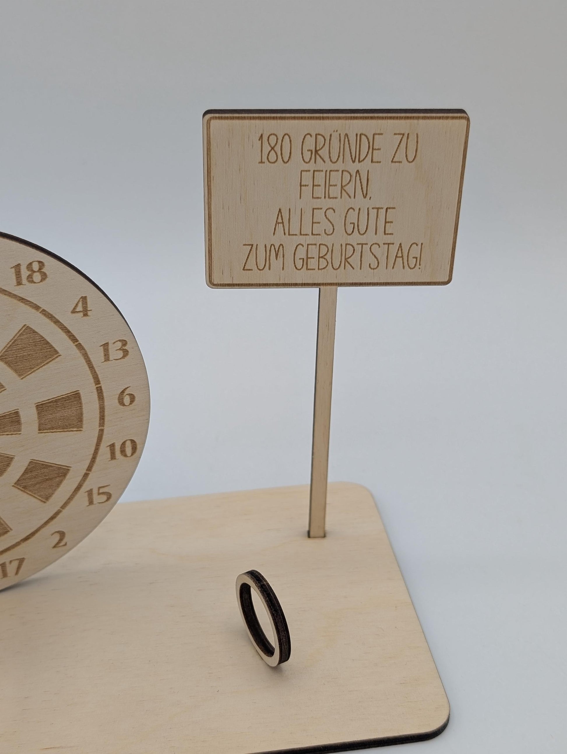 "Personalisierte Dartscheibe aus Holz mit Gravur und Geldhalter, auf einer stabilen Bodenplatte befestigt. Ein Dartpfeil steckt in der Mitte der Scheibe, kann aber auf Wunsch auf eine bestimmte Punktzahl gesetzt werden. Das gravierte Schild zeigt „Happy Birthday – 180 Gründe zu feiern“ oder eine individuelle Widmung. Perfektes Geschenk für Dartspieler zum Geburtstag, Jubiläum oder als Pokal für ein Dartturnier."