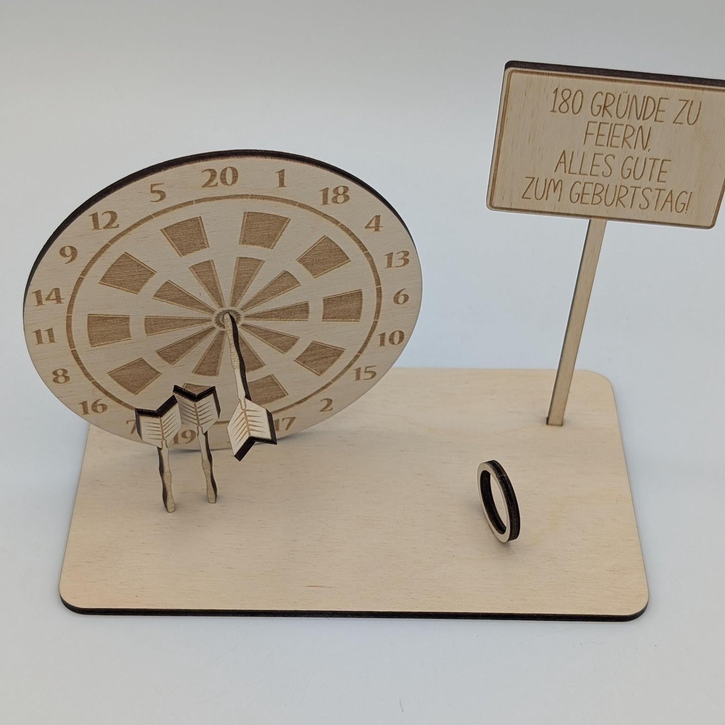 &quot;Personalisierte Dartscheibe aus Holz mit Gravur und Geldhalter, auf einer stabilen Bodenplatte befestigt. Ein Dartpfeil steckt in der Mitte der Scheibe, kann aber auf Wunsch auf eine bestimmte Punktzahl gesetzt werden. Das gravierte Schild zeigt „Happy Birthday – 180 Gründe zu feiern“ oder eine individuelle Widmung. Perfektes Geschenk für Dartspieler zum Geburtstag, Jubiläum oder als Pokal für ein Dartturnier.&quot;